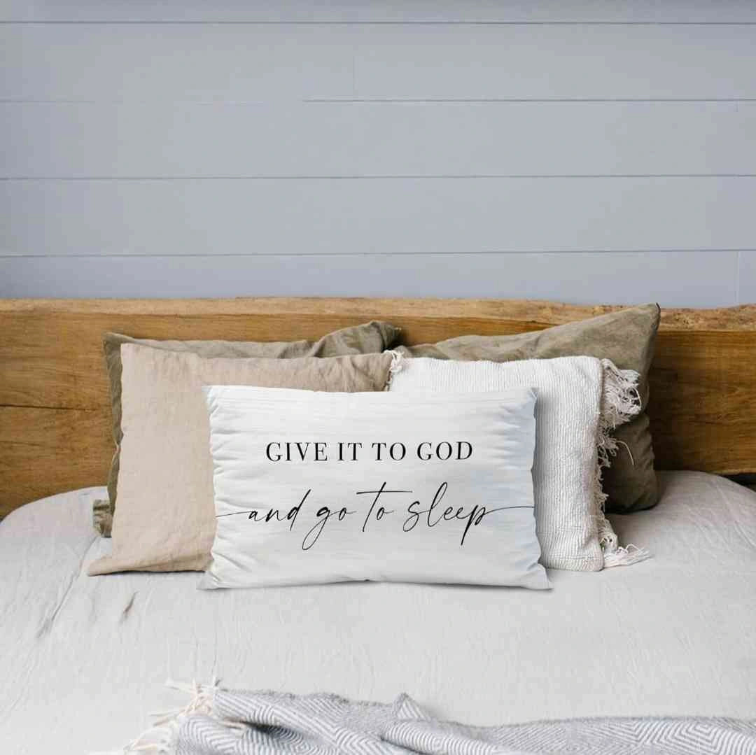 Decorative white pillow with the phrase 'Give It to God and Go to Sleep,' placed on a neutral-toned bed with beige and cream throw pillows, creating a serene bedroom aesthetic.