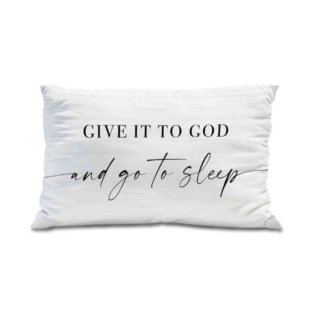 White decorative pillow with the quote 'Give it to God and go to sleep' placed on a minimalist grey bench surrounded by soft, white pillows for a serene look.