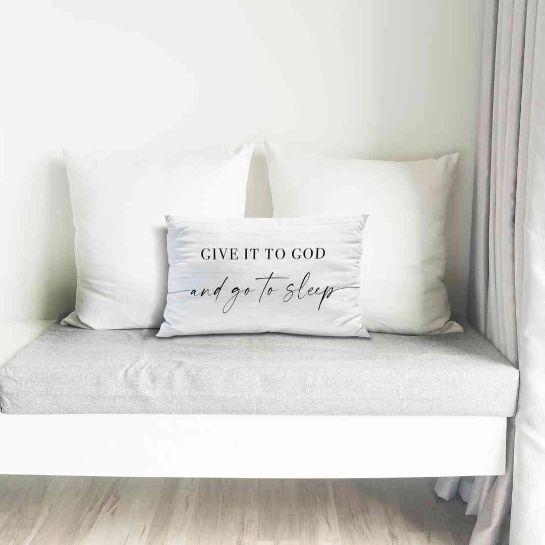 Inspirational white pillow with the quote 'Give It to God and Go to Sleep,' styled on a modern gray bench with white cushions and minimalist decor, perfect for serene spaces.