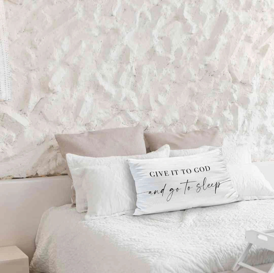 White decorative pillow with the phrase 'Give It to God and Go to Sleep,' placed on a cozy bed with textured white bedding and neutral-toned throw pillows, against a rustic white stone wall.