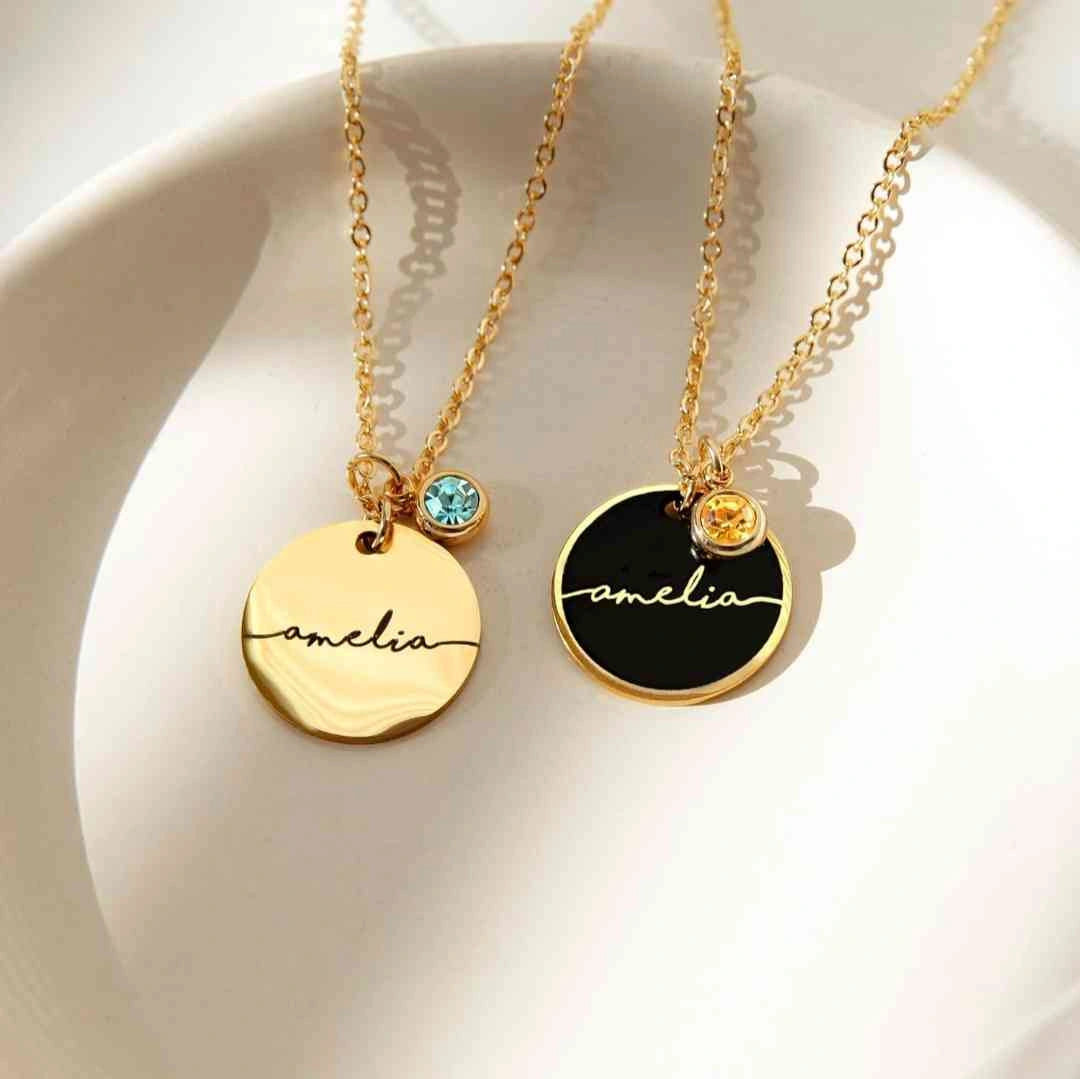 Two gold disc necklaces with custom names and birthstone charms, displayed on a dish.