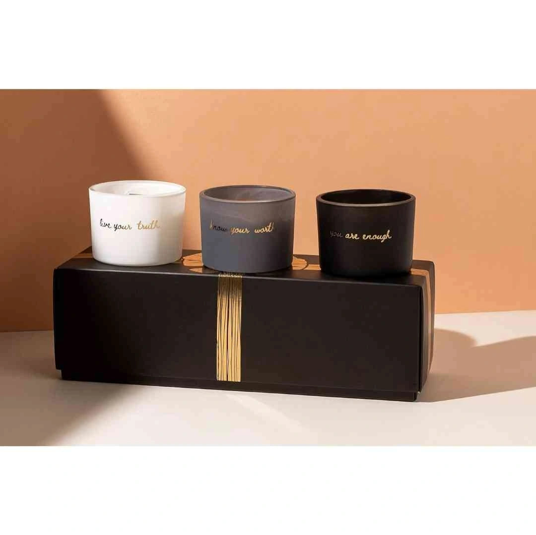 Set of three candles labeled 'Live Your Truth,' 'Know Your Worth,' and 'You Are Enough,' displayed on a sleek black box with gold accents, against a peach-colored background.