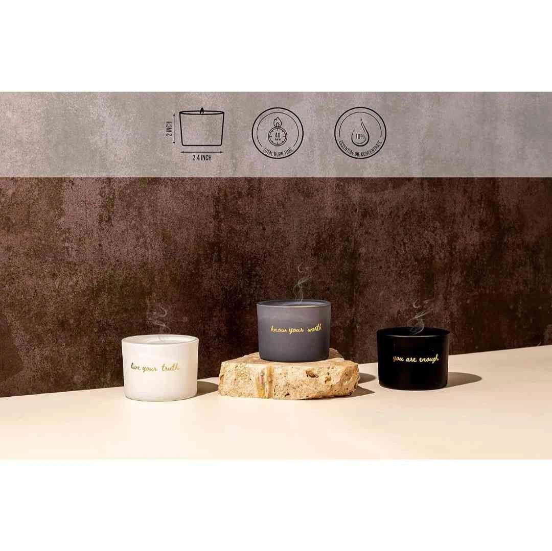 Set of three minimalist candles in black, gray, and white jars with motivational inscriptions, displayed on natural stones against