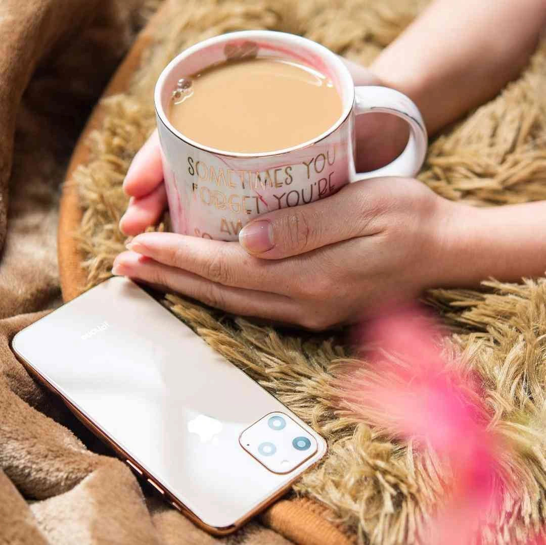 Hands holding a pink marble coffee mug with motivational quote, with a smartphone nearby on a cozy blanket